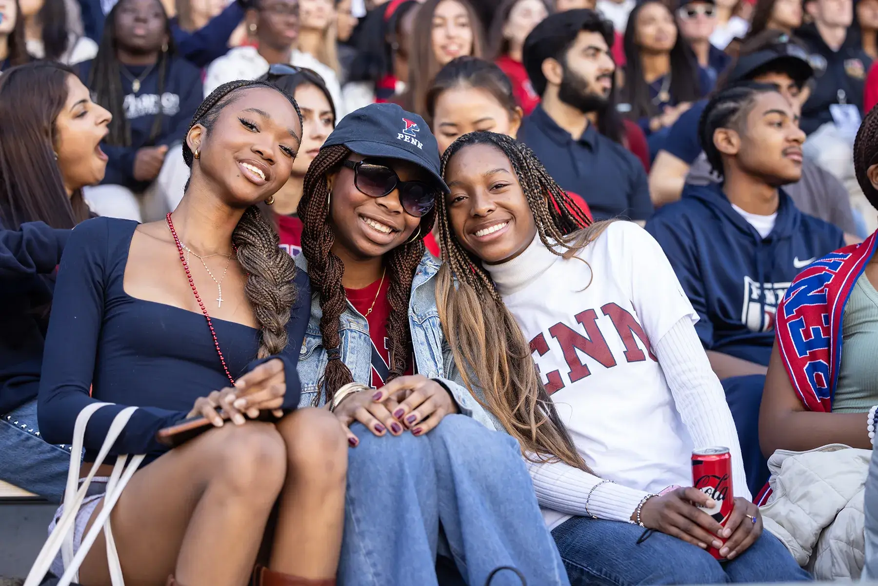 Three smiling Penn students sit together in crowded stadium seating during Homecoming Weekend 2025.