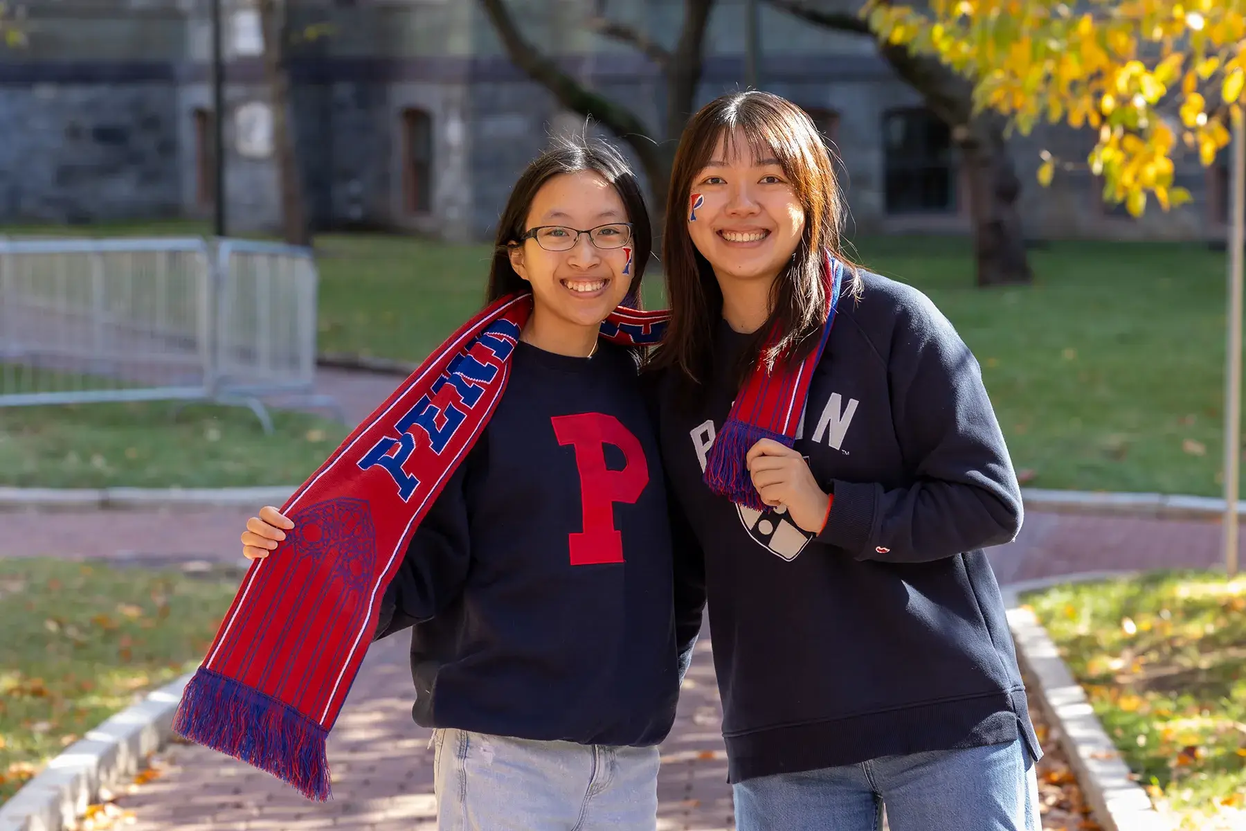 Two smiling Penn students wearing navy sweatshirts, face paint and a red and blue scarf pose outdoors in autumn.