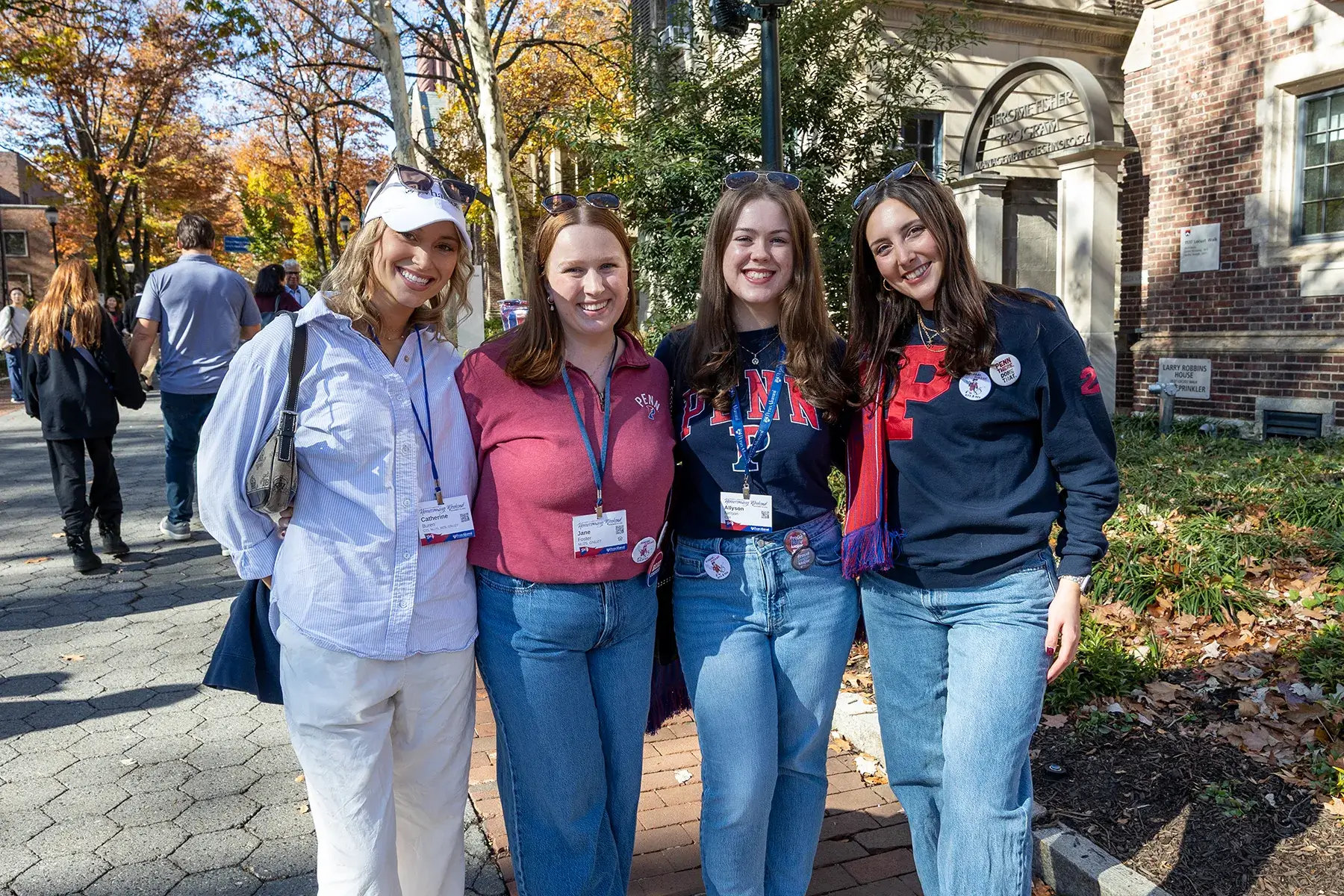 Four Penn Homecoming attendees wearing campus apparel and event badges pose together on a tree-lined walkway.
