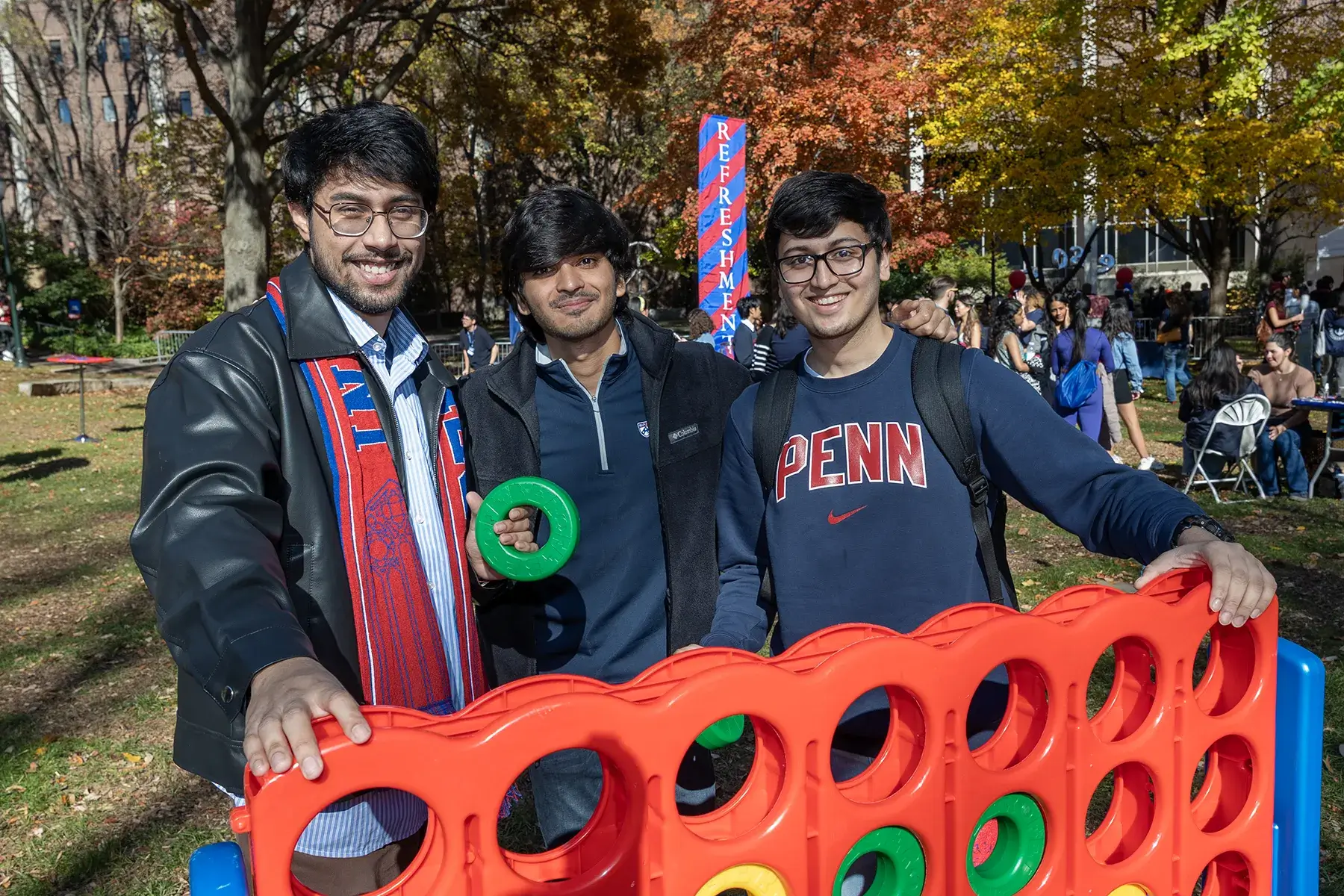 Three Penn Homecoming attendees pose behind a giant four-in-a-row game at an outdoor campus celebration.