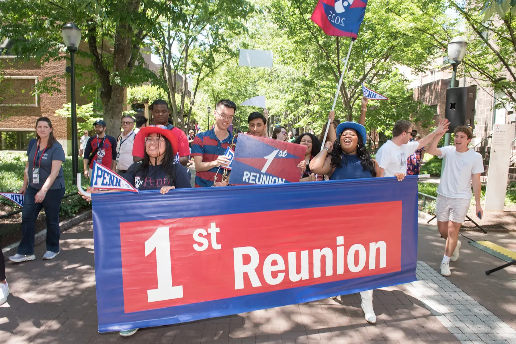 Penn alumni carry a “1st Reunion” banner and wave school pennants during the Alumni Weekend 2026 parade on campus.