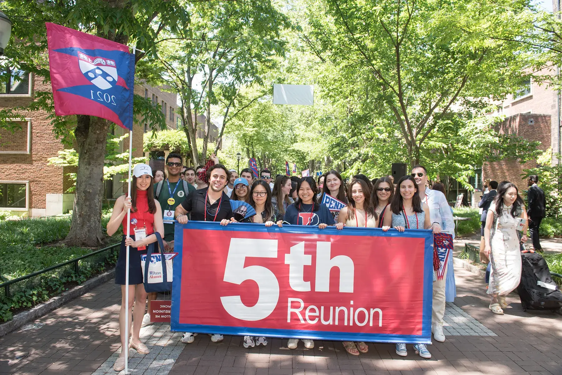 Penn alumni gather behind a “5th Reunion” banner beneath leafy trees during Alumni Weekend 2026.
