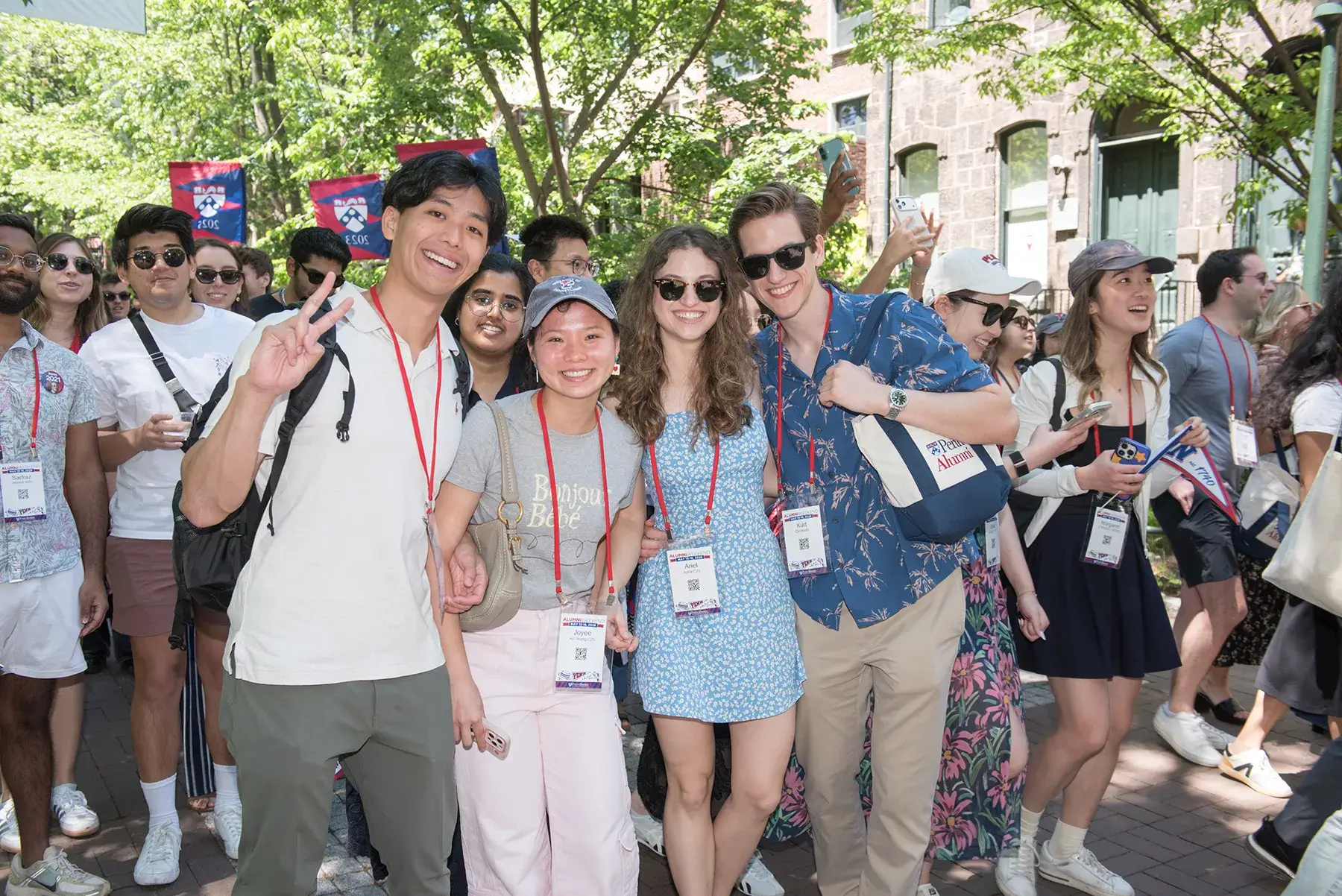 Four Penn alumni wearing reunion badges pose together amid a crowded outdoor Alumni Weekend 2026 celebration.