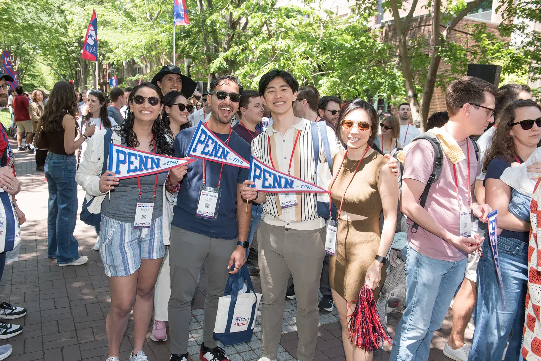 Four alumni hold Penn pennants and smile along a tree-lined campus walkway during Alumni Weekend 2026.