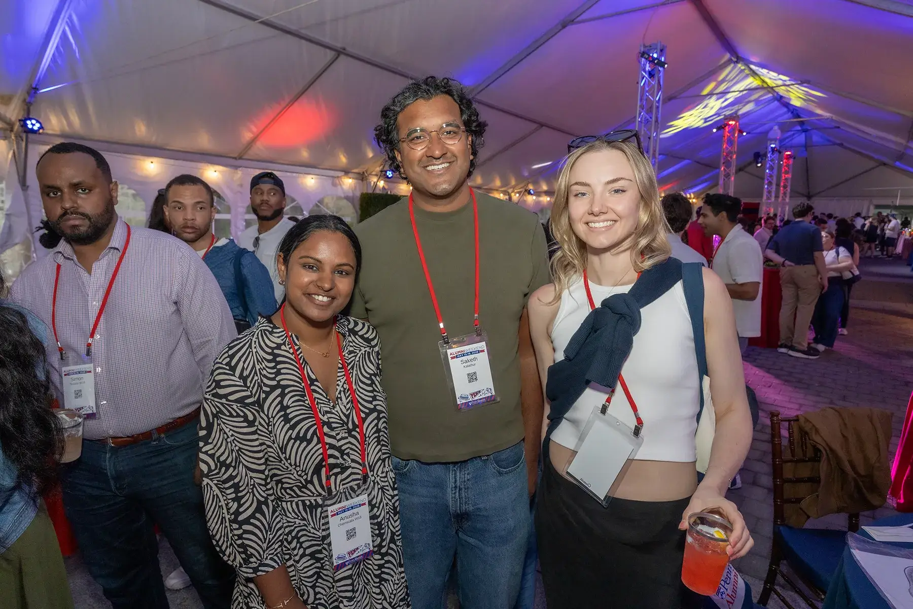 Three alumni wearing event badges pose inside a tent lit with red, blue, and purple lights during Alumni Weekend 2026.