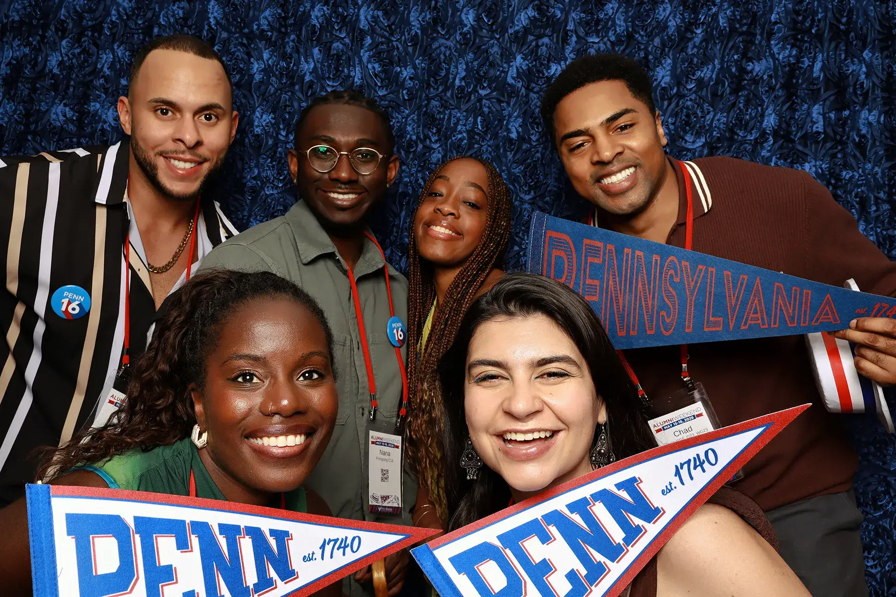 Six Penn alumni smile for a photo booth picture while holding Pennsylvania and Penn pennants during Alumni Weekend 2026.