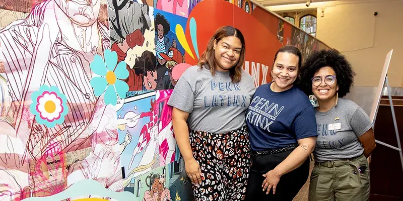 Three people wearing Penn Latinx shirts stand together beside a colorful mural.