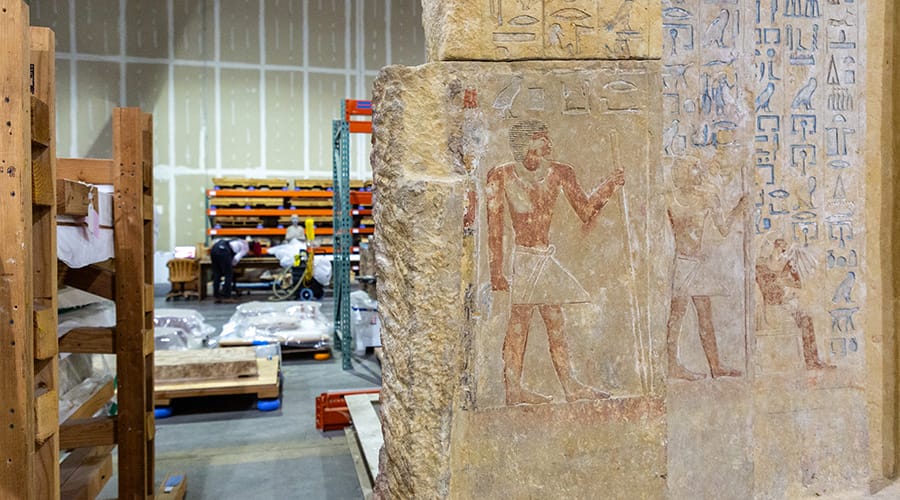 An ancient Egyptian relief, richly adorned with hieroglyphs and depictions of figures in traditional attire, is temporarily stored in a modern warehouse setting. The relief features a prominent figure in a standing pose, holding a staff, and is flanked by smaller detailed carvings. The background includes a visible storage area with wooden pallets and industrial shelves, filled with various items under protective covers.