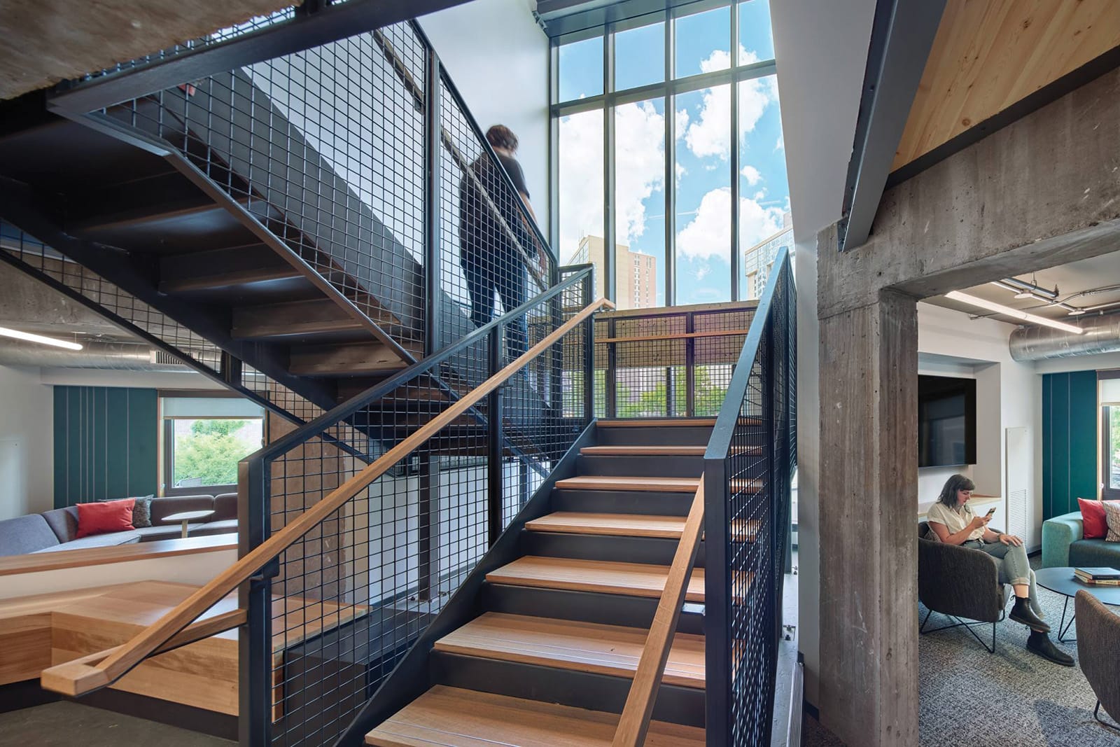 Interior view of a modern building with a stylish wooden staircase enclosed by a metal mesh grid, connecting two levels. The area is bathed in natural light from large windows showing an urban landscape. To the right, a cozy seating area with an individual reading adds to the inviting ambiance of the space.