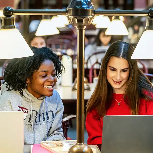 Close-up of two smiling Penn students studying together at a table with laptops and library lamps.