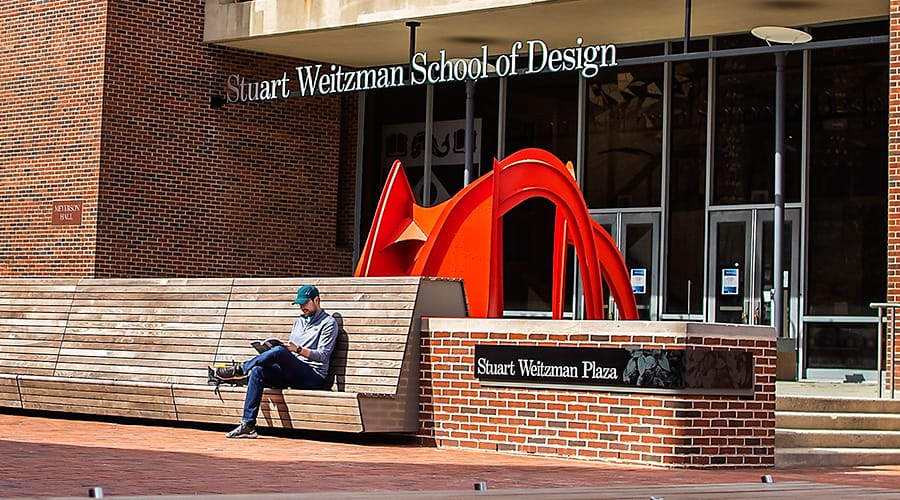 A view of Meyerson Hall at the University of Pennsylvania. In the foreground, a person in casual attire sits alone on a bench, reading a magazine.